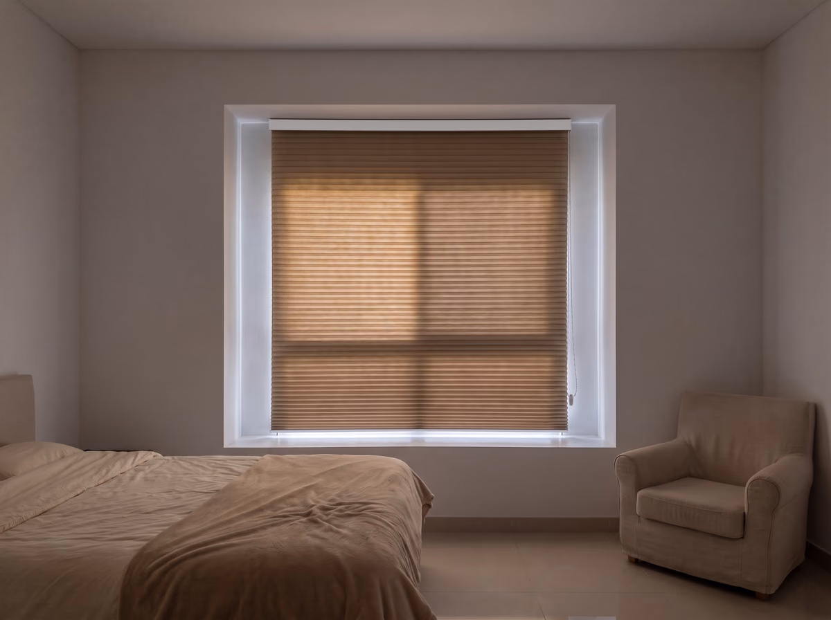 Villa bedroom at midday with a taupe dimout honeycomb blind lowered inside the recess, a soft glow through it and the room in shade
