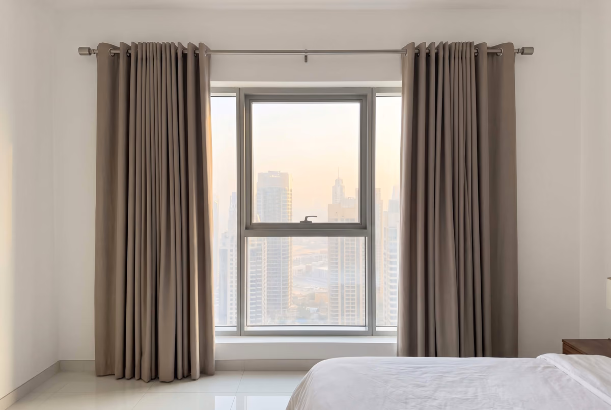 Bedroom with taupe eyelet curtains on a brushed-steel pole, drawn back either side of a tall window over Downtown Dubai towers