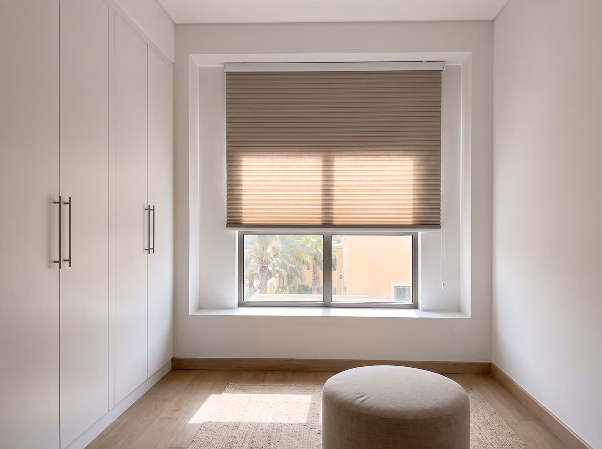 Dressing room with a taupe honeycomb blind lowered two-thirds inside the window recess, white wardrobes and a linen ottoman in front