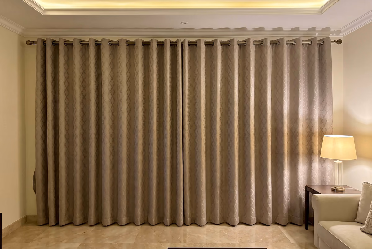 Taupe jacquard eyelet curtains closed across a living room wall in the evening, on an antique brass pole under a cove-lit ceiling