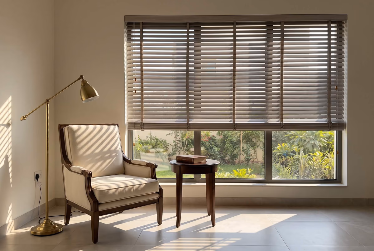 Taupe wooden blind raised halfway over a wide window onto a garden, an armchair, a side table and a brass floor lamp in striped sunlight