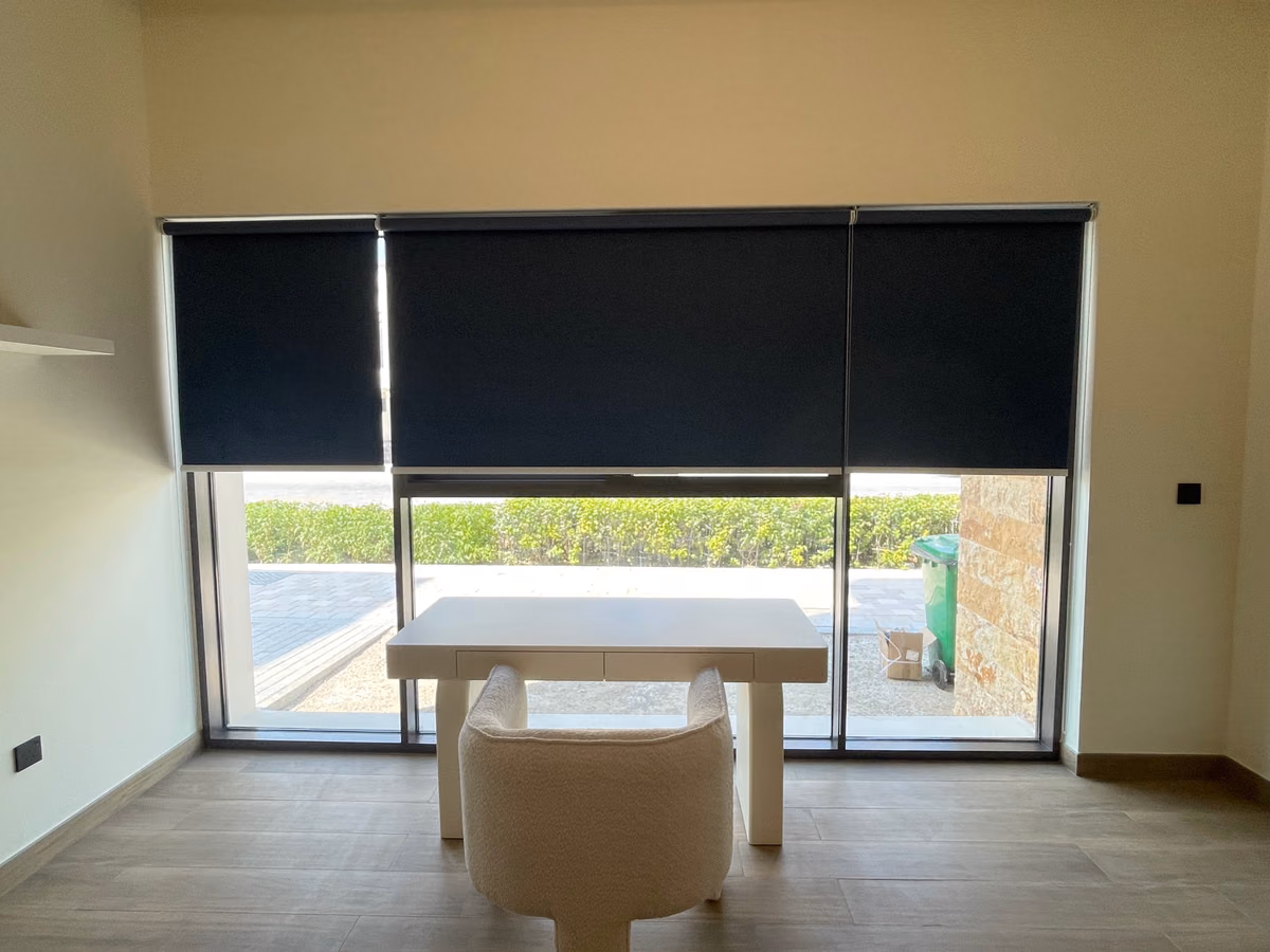 Three dark navy roller blinds half lowered across a ground-floor study window, a white desk and chair in front and a hedge outside
