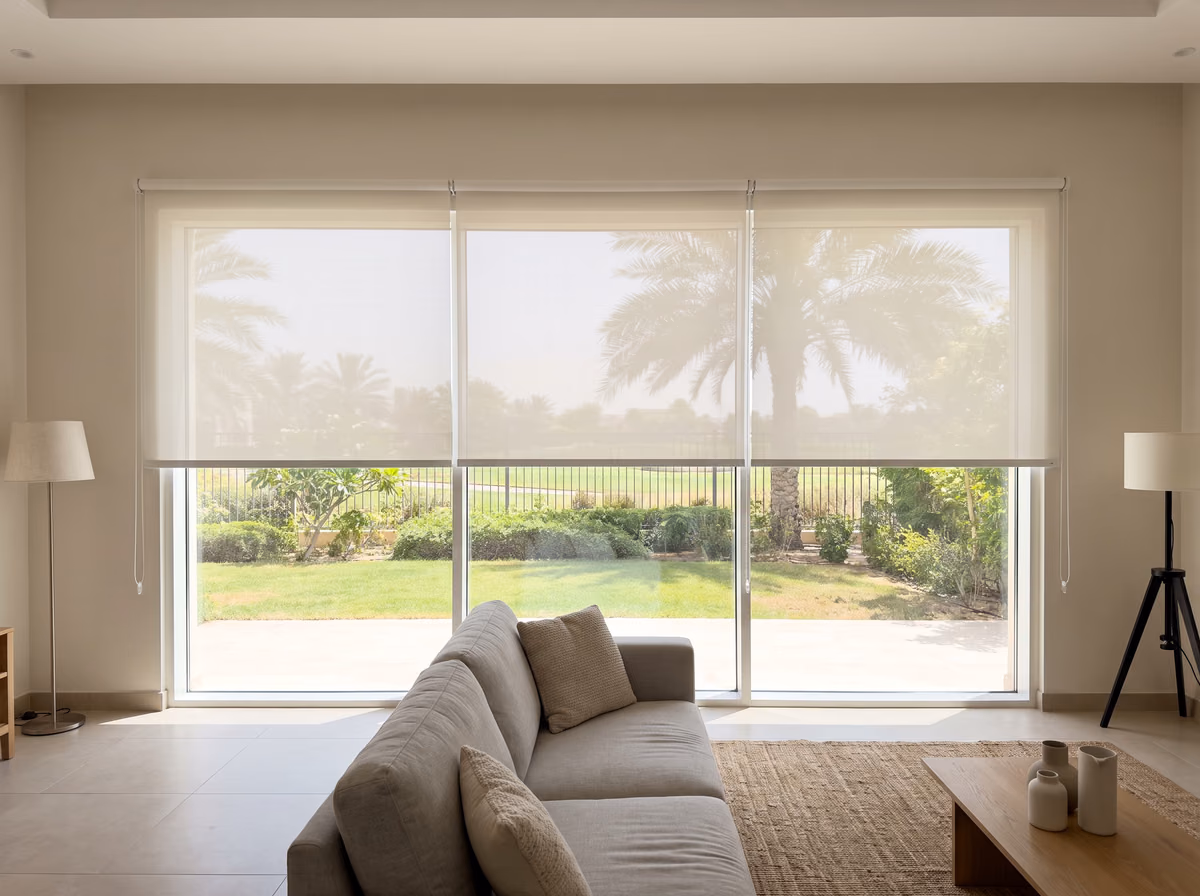 Villa living room with three white sunscreen roller blinds over wide glass doors, lowered to one height, palms and a garden beyond