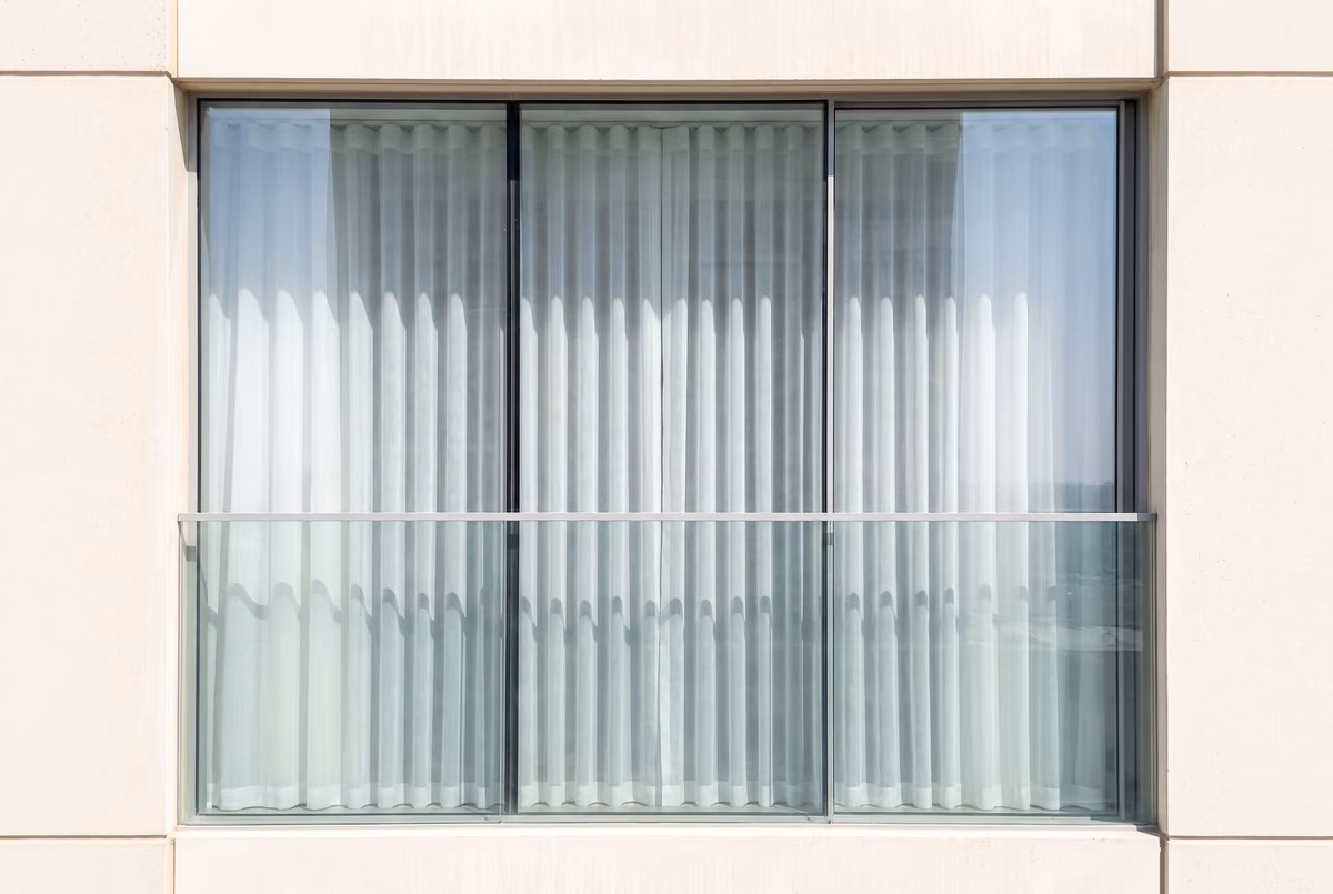 Tower bedroom window seen from the building opposite at midday, a white wave sheer closed behind the glass and nothing inside visible