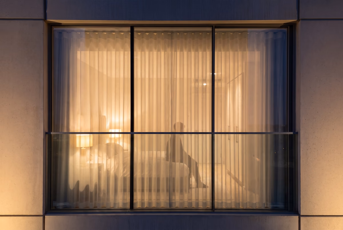 The same tower bedroom window at night with the lamps on, the closed sheer showing a bed, bedside lamps, a wardrobe and a person sitting on the bed