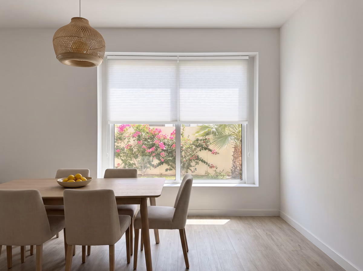Dining room with two white honeycomb blinds side by side inside the window recess, half lowered, bougainvillea and a palm beyond