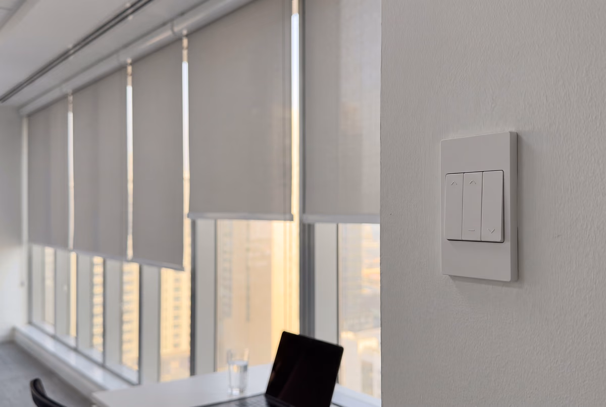 Slim white wall switch with up and down arrows beside a row of light grey roller blinds lowered over office windows