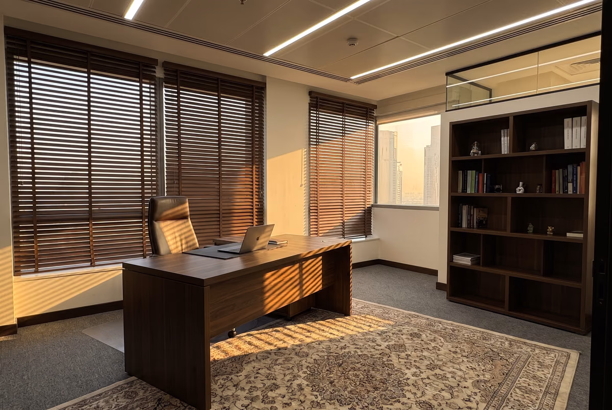 Walnut-colour faux wood blinds tilted half open in an executive office, low sun throwing stripes across the desk and rug