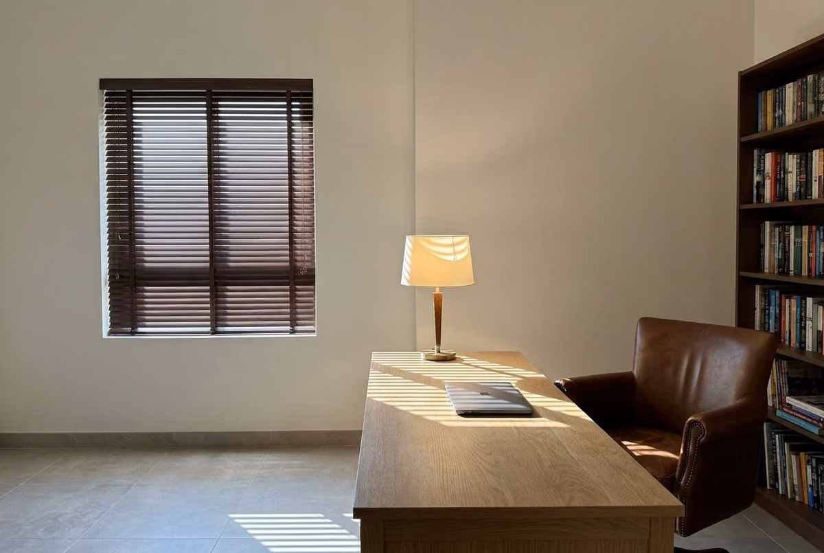 Study with a dark walnut wooden blind on ladder tapes, lowered inside the window recess, a lamp lit on an oak desk and a leather chair beside a bookcase