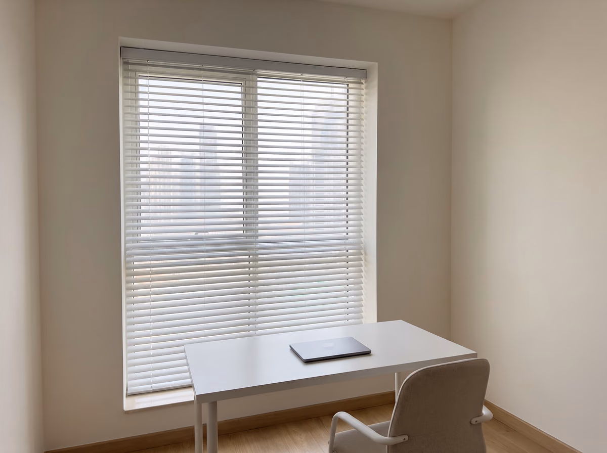 White venetian blind tilted half open inside a study window recess, a closed laptop on a white desk in front