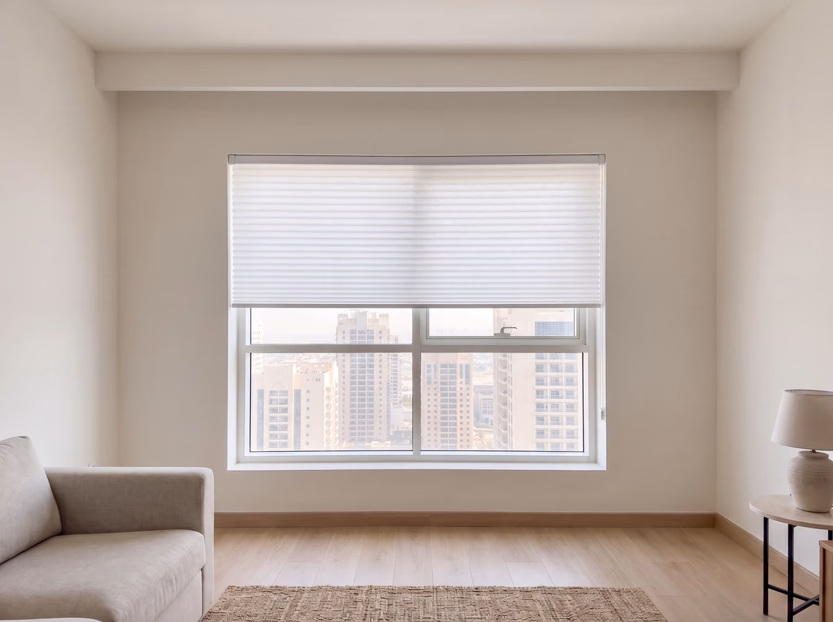 White honeycomb blind lowered halfway inside a living room window recess, daylight glowing through its pleats and the towers clear below