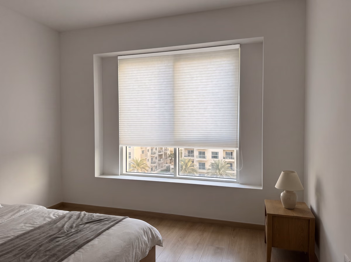 Bedroom window with a white honeycomb blind inside the recess, lowered three-quarters, a bead chain at its right end and low-rise blocks below