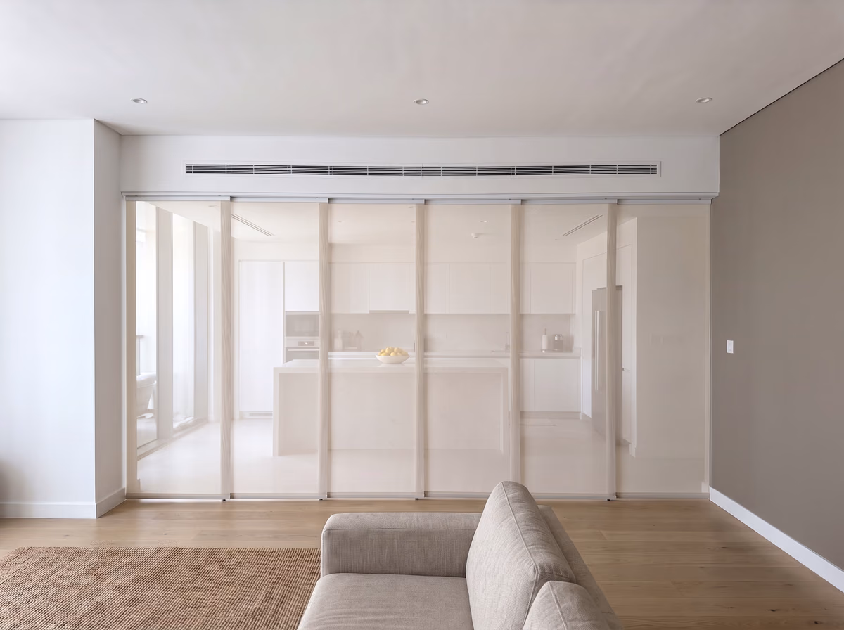 White sheer panel blinds hung from a ceiling track and drawn across a wide glass partition, an open-plan kitchen softened behind them