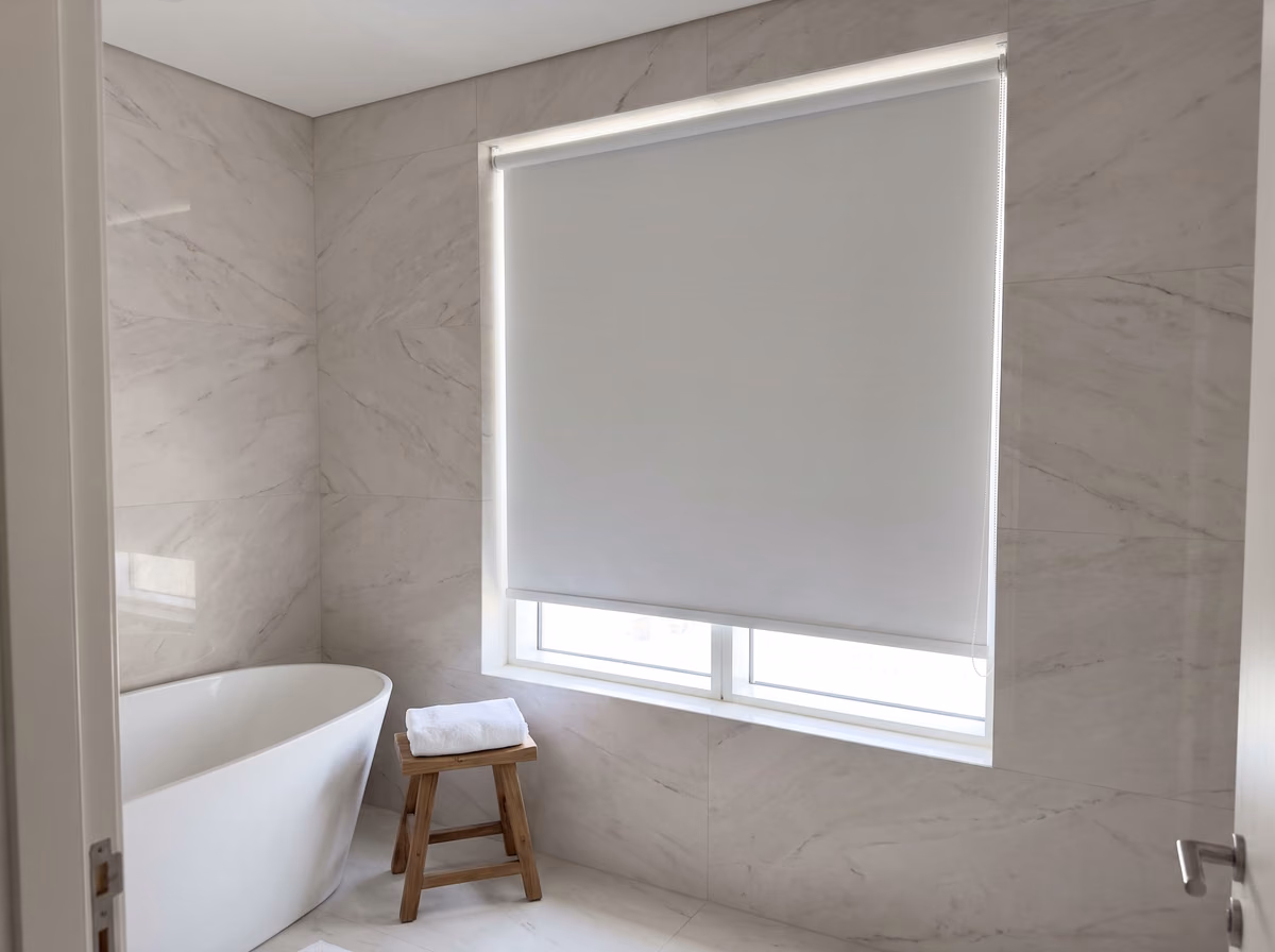 Marble bathroom with a plain white roller blind lowered most of the way over the window beside a freestanding bath, a towel on a wooden stool