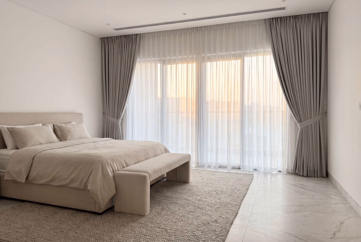 White sheer drawn across a bedroom's glass doors, grey pinch-pleat curtains tied back at each side and a bench at the foot of the bed