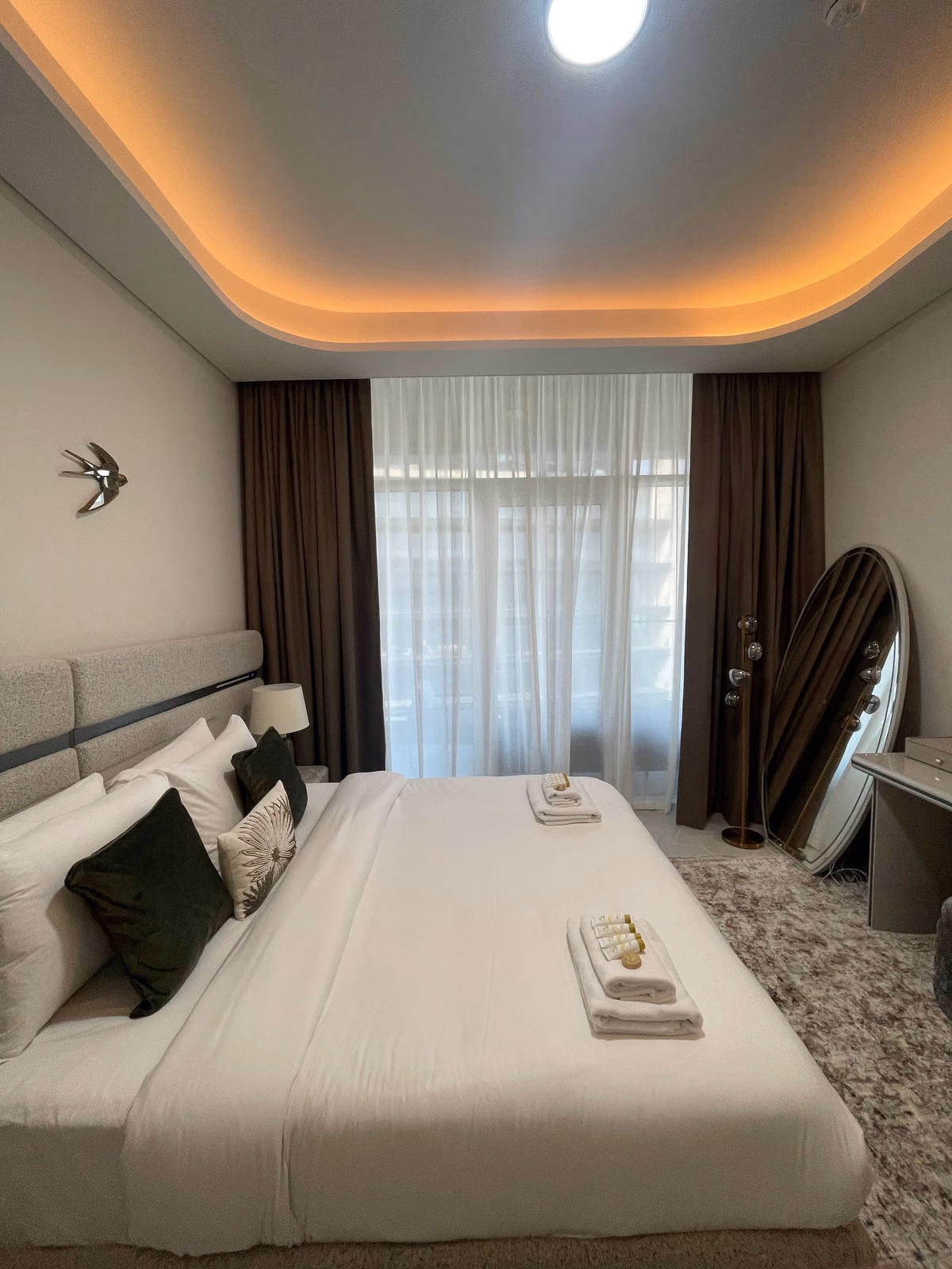 A white voile sheer drawn across a bedroom window between two brown blackout curtains stacked at the sides, a bed and an oval floor mirror in front