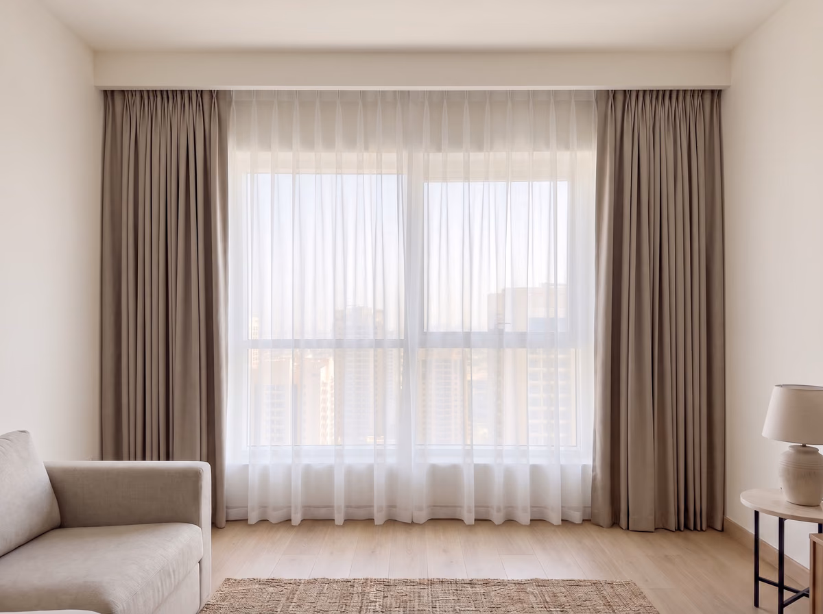 The same window with a white voile sheer drawn across the glass and taupe blackout curtains stacked at each side, both from the ceiling pocket