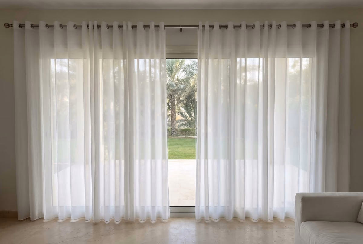 White sheer eyelet curtains on a silver pole across villa patio doors, parted in the middle onto a lawn and a palm tree