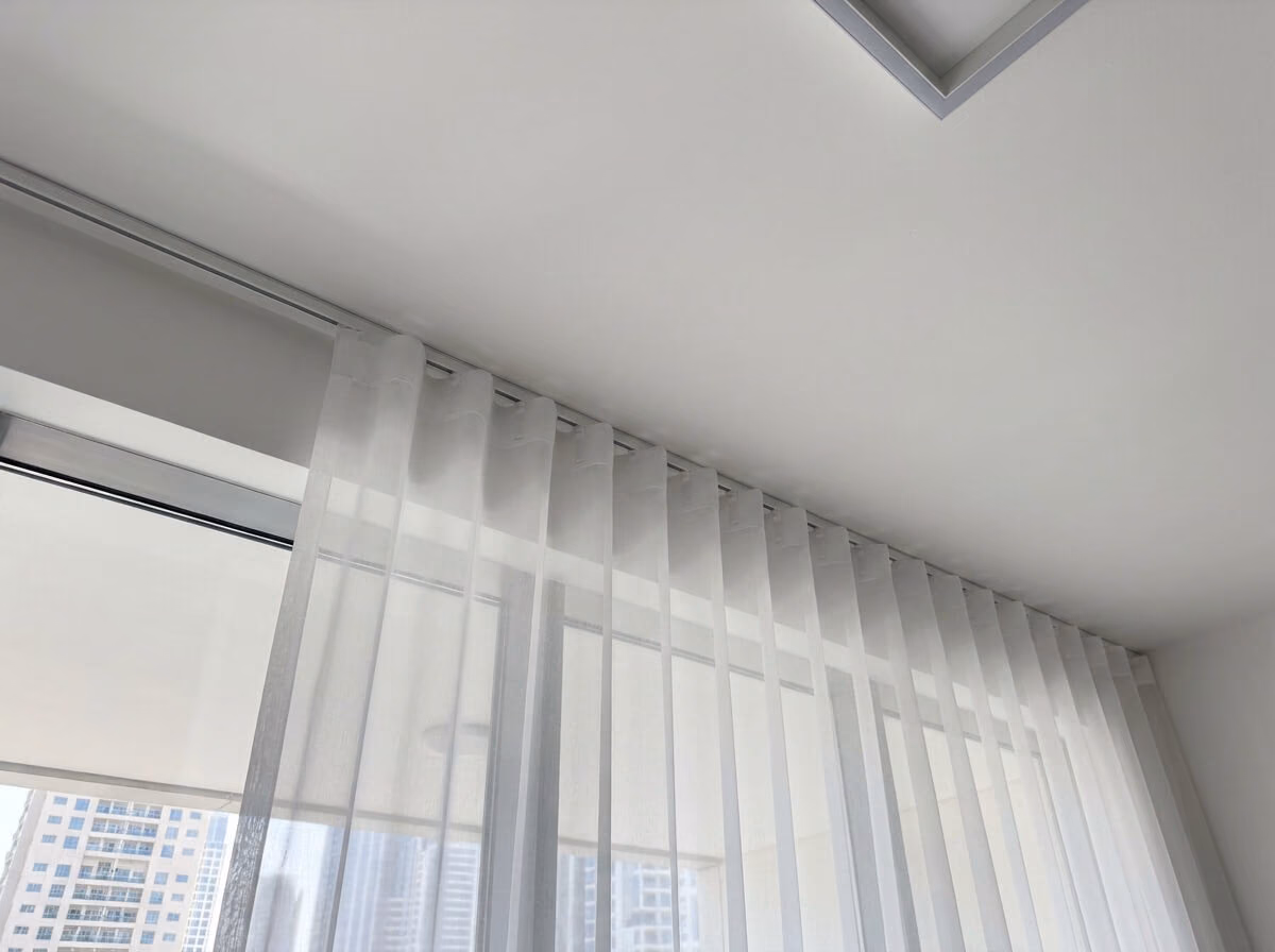 Close-up of a white sheer wave curtain clipped to carriers in a white ceiling track, falling in even folds, towers outside