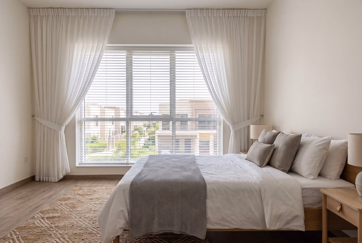 White sheer curtains tied back either side of a bedroom window, a white slatted blind lowered behind them