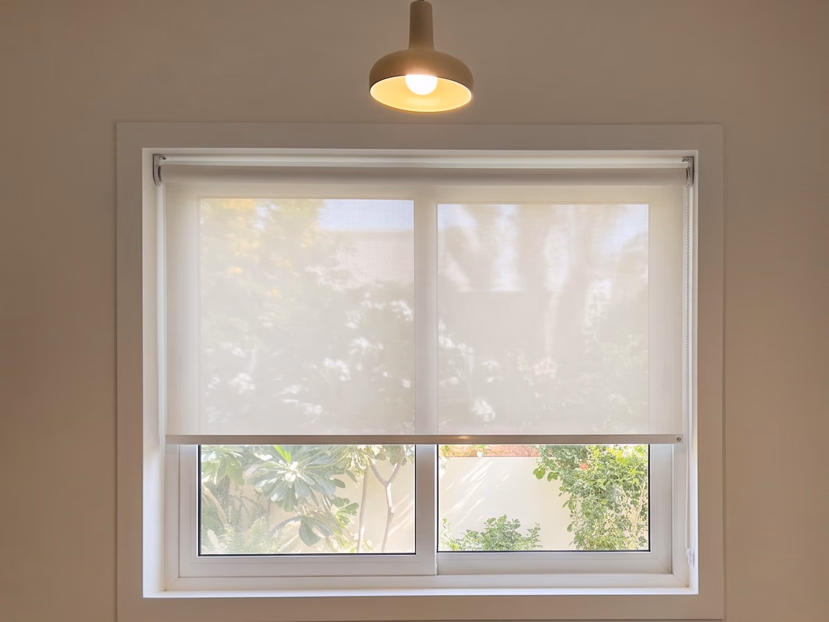 White sunscreen roller blind with a bead chain at its side, lowered most of the way inside a kitchen window, a pendant light above