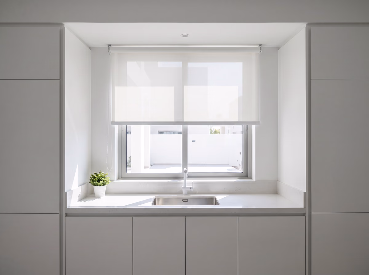 White kitchen with a white sunscreen roller blind lowered halfway in the window above a stone counter and a steel sink, daylight softened through it