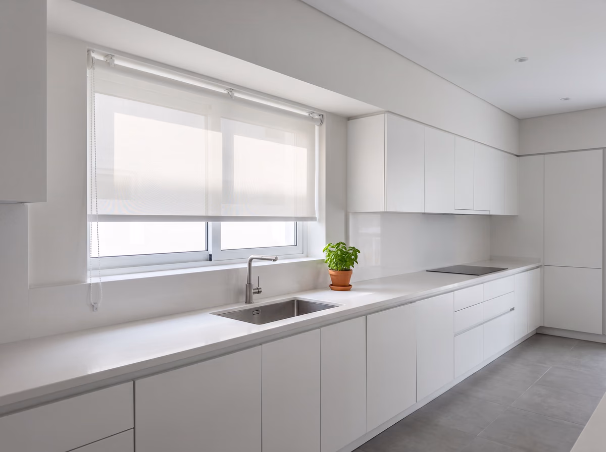 Kitchen window with a white sunscreen roller blind lowered part way inside the recess, its bead chain held taut by a clip on the wall