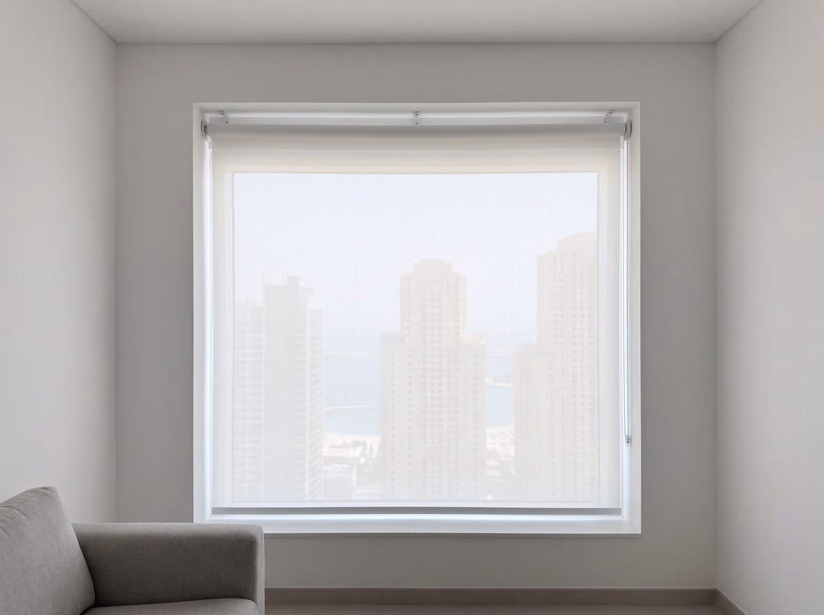 White sunscreen roller blind fully lowered over a tower window at midday, the towers and the sea pale but visible through the fabric