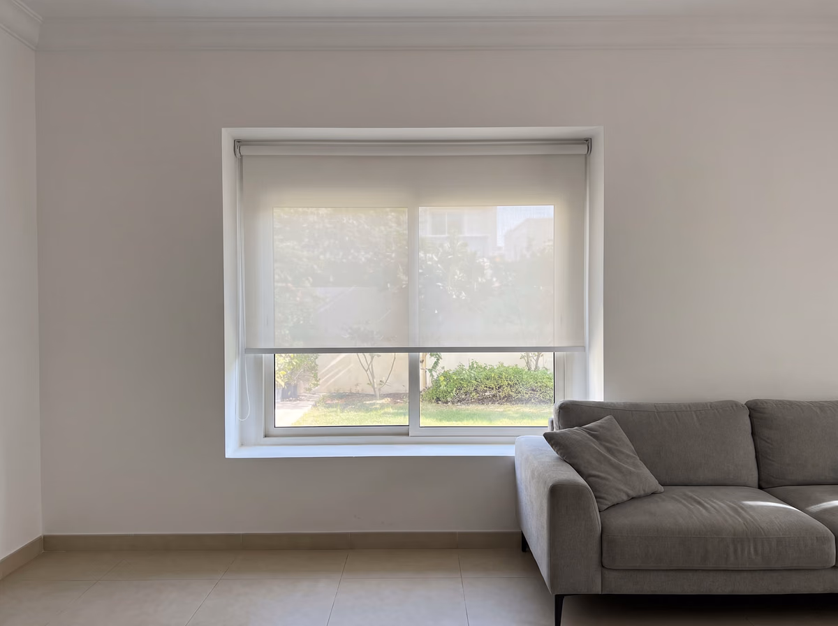 Villa living room with a white sunscreen roller blind lowered part way inside the window recess, garden plants softly through it