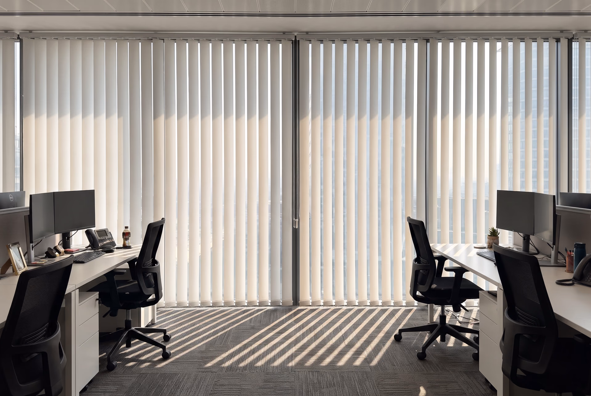 Open-plan tower office with white sunscreen vertical blinds drawn across floor-to-ceiling glass, sun striping the carpet between rows of desks