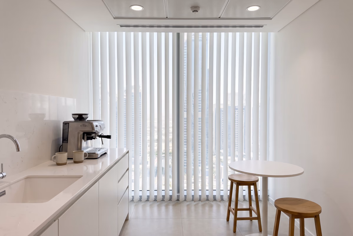 White vertical blinds closed across a tall window in an office pantry with a sink, a coffee machine and two stools