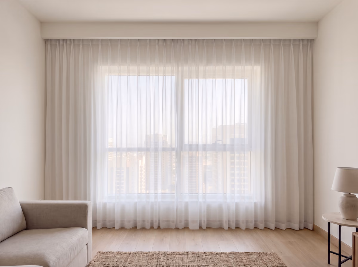 Living room window with white voile sheer curtains in a pinch-pleat heading drawn closed to the floor, the towers soft behind the weave