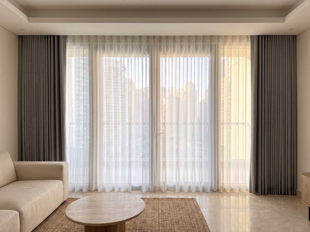 Tower living room with a white ripple-fold sheer drawn across floor-to-ceiling glass and grey curtains stacked at each end, towers beyond