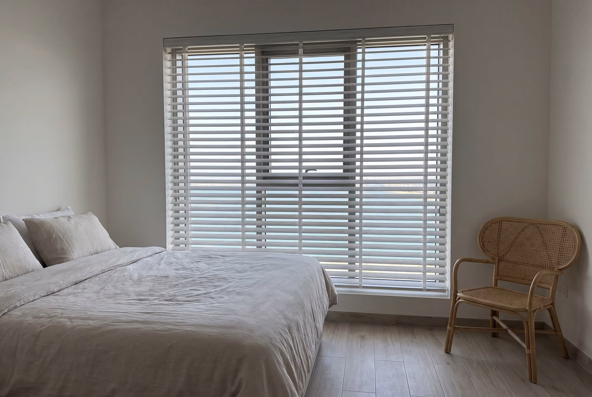 Tower bedroom with a white wooden blind lowered across a wide window, the slats tilted open onto the sea and the coastline, a rattan chair in the corner