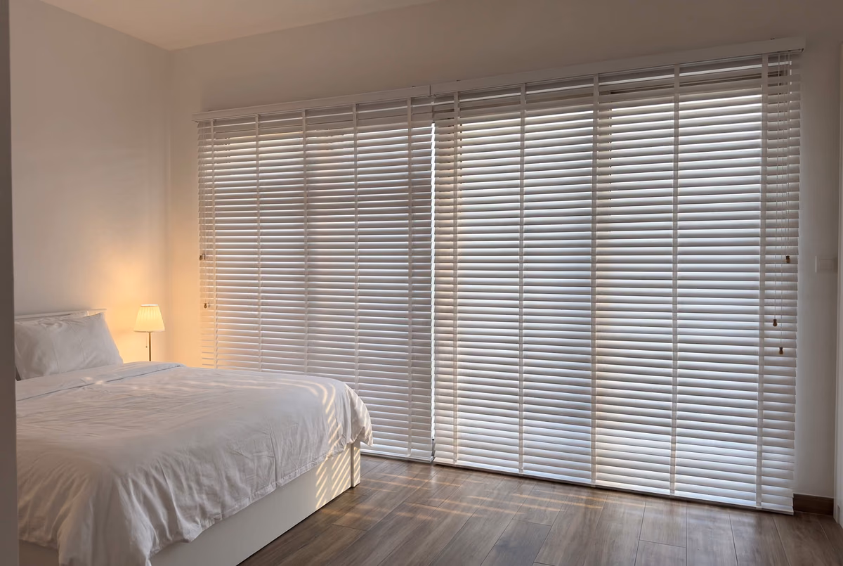 Bedroom with white wooden blinds closed across a wall of glass, daylight glowing between the slats onto a wood floor and a lamp lit beside the bed