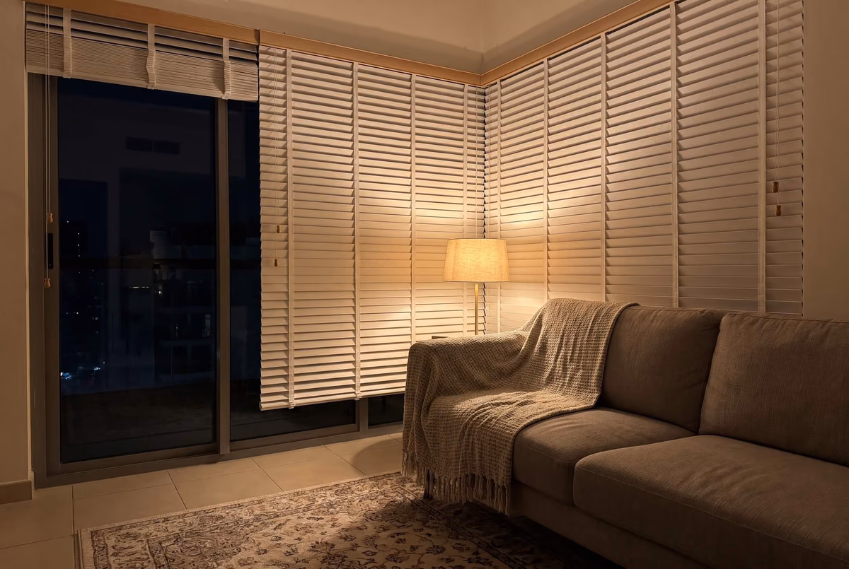 Apartment living room at night with white wooden blinds closed along two walls of corner glass, a floor lamp glowing against them and a blind raised over the balcony door