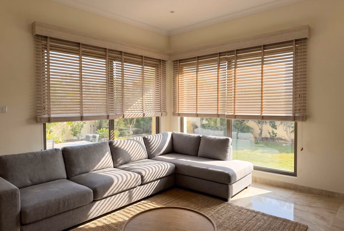 Villa living room with whitewashed wooden blinds under matching pelmets, raised partway on two corner windows above a grey sofa, a garden beyond